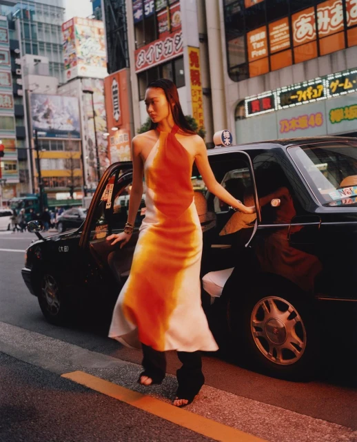 A woman in a halter dress stands by a black taxi in a city street with tall buildings and bright signage, creating a fashion editorial.