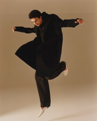 Full-length studio shot of a young adult man leaping mid-air wearing a black coat and pants with white shoes, against a beige backdrop.