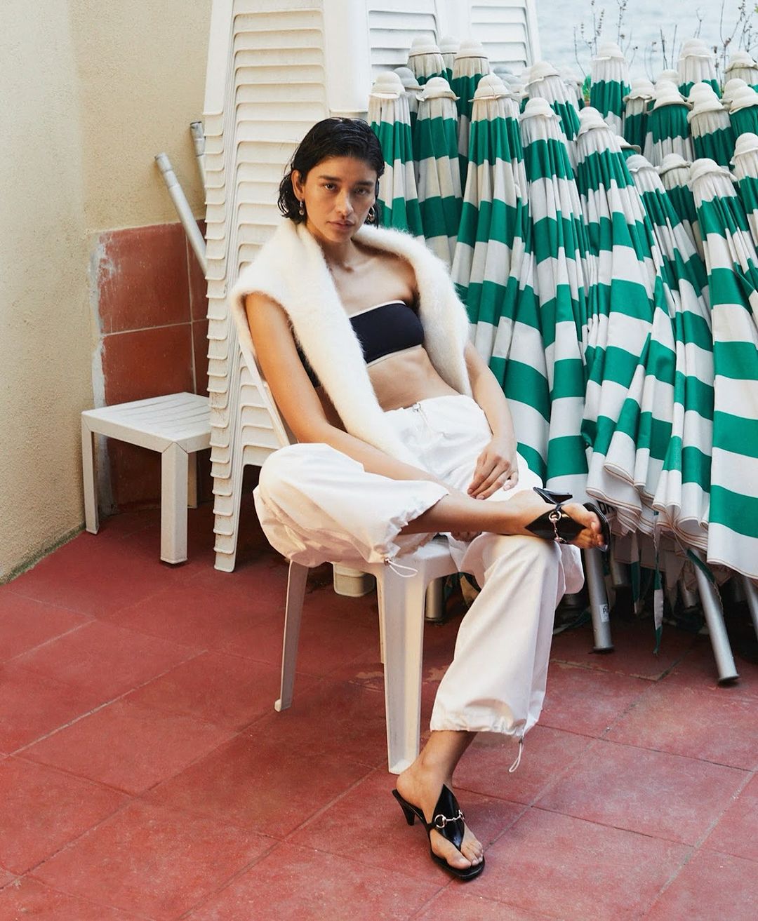 A woman in a black bandeau top and white pants poses seated on a white chair outside, with stacked beach umbrellas behind her.