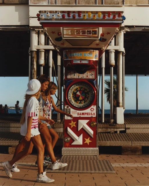Three young women walk on a sidewalk past a vintage amusement game, under the sunlight, with the ocean visible in the background.