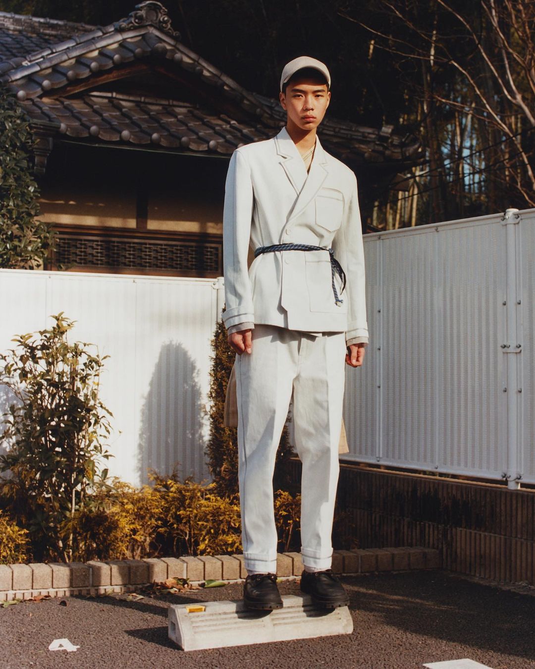 A young adult male model stands in a grey suit and cap in an outdoor fashion editorial shoot with a Japanese house background.