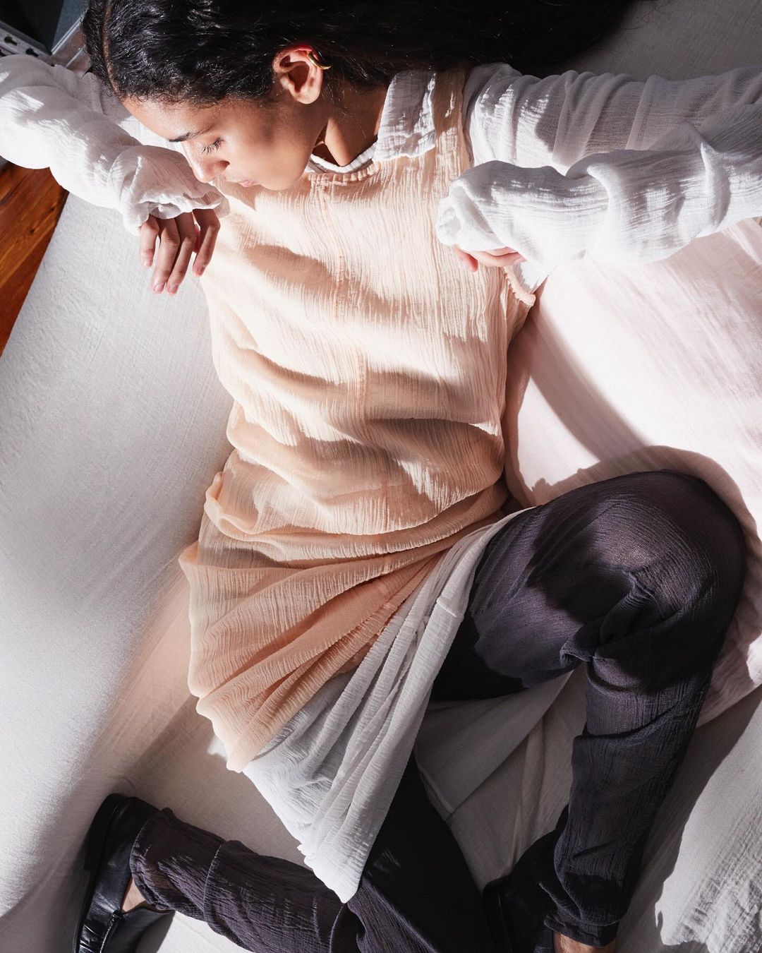 Model wearing pale pink sleeveless dress over white blouse and dark trousers resting on white sofa