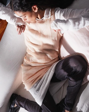 Model wearing pale pink sleeveless dress over white blouse and dark trousers resting on white sofa