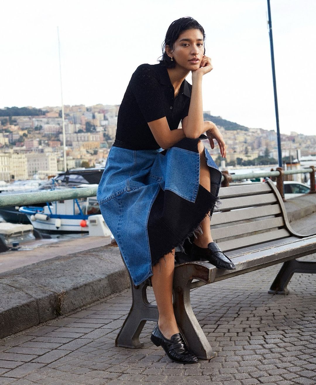 Adult woman poses on a bench in outdoor urban setting with city and harbor in background. She wears a blue and black skirt and black top.