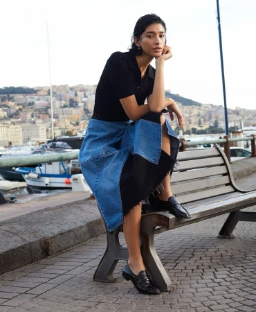 Adult woman poses on a bench in outdoor urban setting with city and harbor in background. She wears a blue and black skirt and black top.