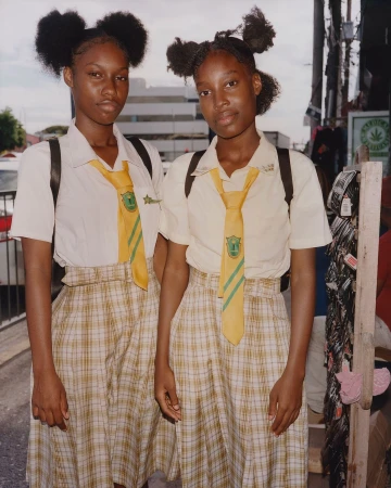 Two young Black women in school uniforms pose outdoors with serious expressions, captured in a medium shot on a city street in daylight.