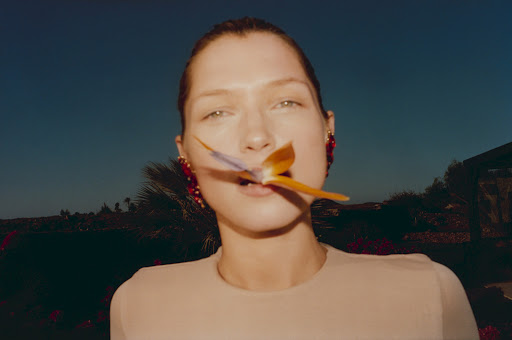 Close-up of a woman with an orchid in her mouth against a dark blue sky, wearing statement red earrings and a tan-colored top.