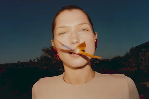 Close-up of a woman with an orchid in her mouth against a dark blue sky, wearing statement red earrings and a tan-colored top.