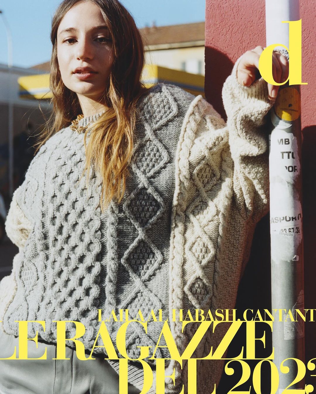 A woman in a patterned knit sweater stands in front of a building on a sunny day for a fashion editorial, creating a stylish visual.