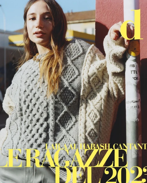 A woman in a patterned knit sweater stands in front of a building on a sunny day for a fashion editorial, creating a stylish visual.