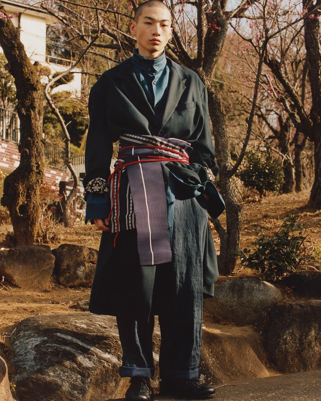 Asian man poses in outdoor fashion shot wearing oversized black coat, blue shirt, wide dark pants, and decorative sash in natural light.