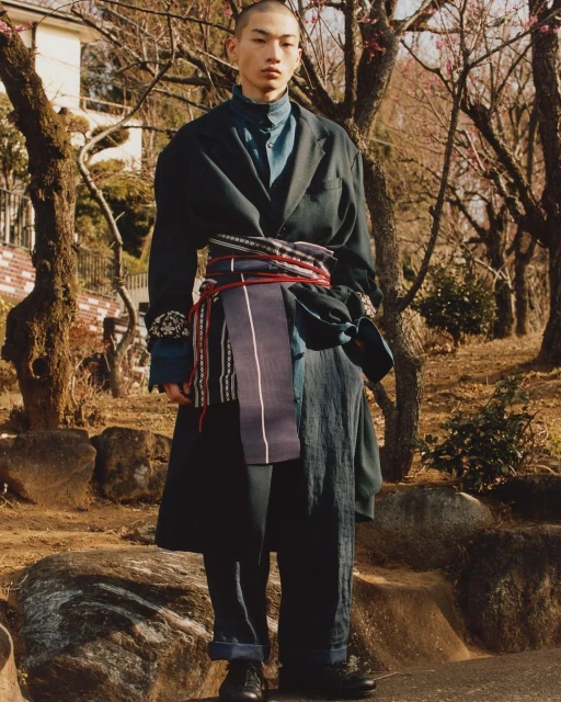 Asian man poses in outdoor fashion shot wearing oversized black coat, blue shirt, wide dark pants, and decorative sash in natural light.