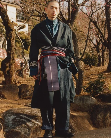 Asian man poses in outdoor fashion shot wearing oversized black coat, blue shirt, wide dark pants, and decorative sash in natural light.