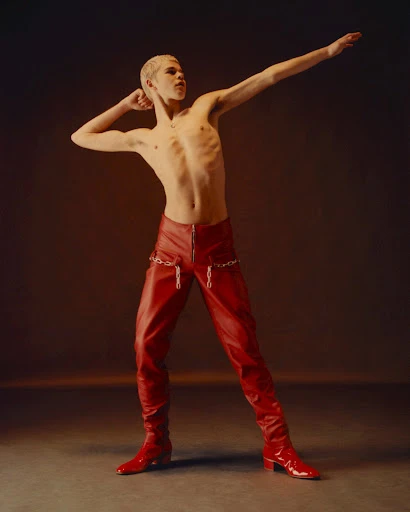 Shirtless person with platinum buzzcut, in red leather pants and matching boots, poses with arms raised in a studio setting.