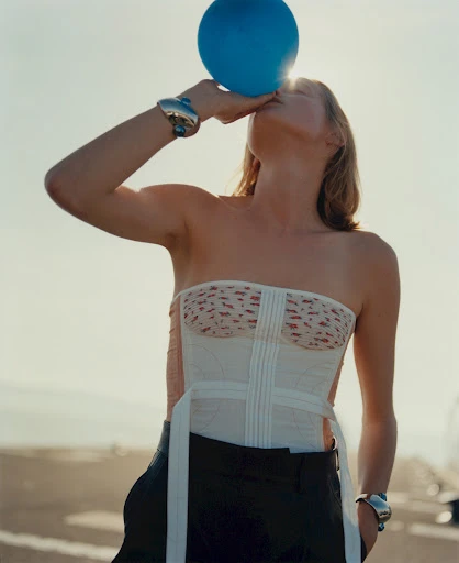 Adult female in corset and dark pants blowing up a blue balloon against a bright, clear sky, accessorized with metallic bangles.
