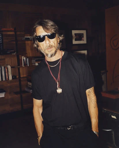 Portrait of a man wearing sunglasses and a black t-shirt, posing in an indoor setting with a bookshelf and artwork in the background.