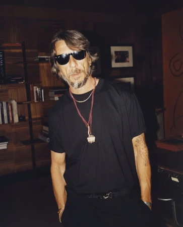 Portrait of a man wearing sunglasses and a black t-shirt, posing in an indoor setting with a bookshelf and artwork in the background.
