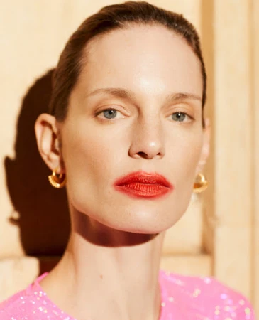 Close-up portrait of a woman wearing gold earrings and a pink top, photographed by Paolo Zerbini with hard lighting casting shadows.