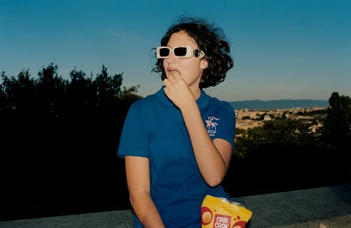 Young woman with sunglasses eating chips outdoors against a cityscape backdrop during daytime, wearing a blue polo shirt.