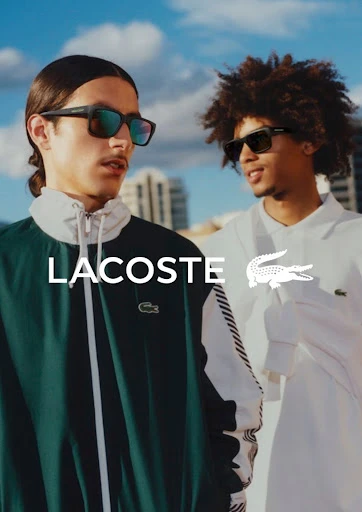 Two adult males wear Lacoste clothing and sunglasses in an outdoor setting with a modern building and blue sky in the background.