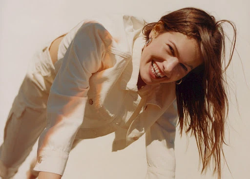Smiling woman wearing an off-white denim jacket leaning forward against a plain light background