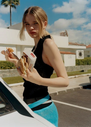 Young woman in a black top is standing outside eating a burger, fries, and drink with a building and palm tree in background.