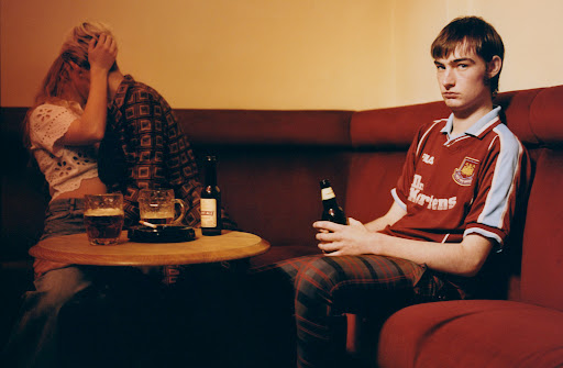 Indoor scene shows a forlorn young man on a red couch with a beer, while a couple kiss at a table with beer mugs.