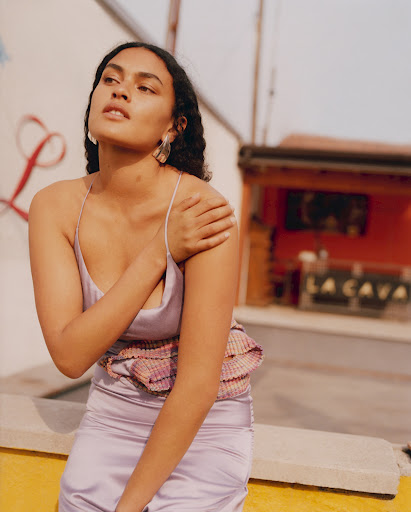 A woman with long dark hair poses in a lilac satin dress outdoors in natural light, with a building in the background.