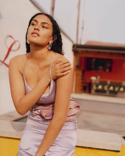 A woman with long dark hair poses in a lilac satin dress outdoors in natural light, with a building in the background.