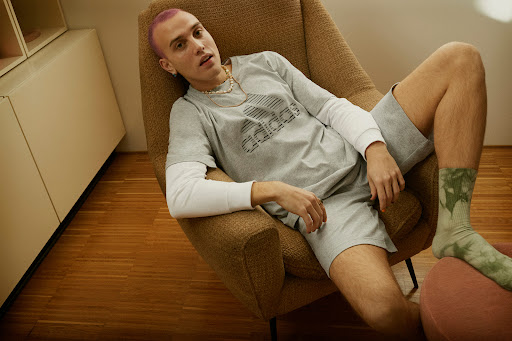 Man in Adidas t-shirt and shorts lounges in armchair. Interior shot with wood floors and muted colors. Fashion photography.