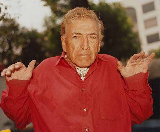 Portrait of an elderly Caucasian man with a red shirt and silver pendant necklace outdoors with trees and a building in the background.