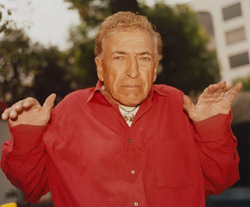 Portrait of an elderly Caucasian man with a red shirt and silver pendant necklace outdoors with trees and a building in the background.