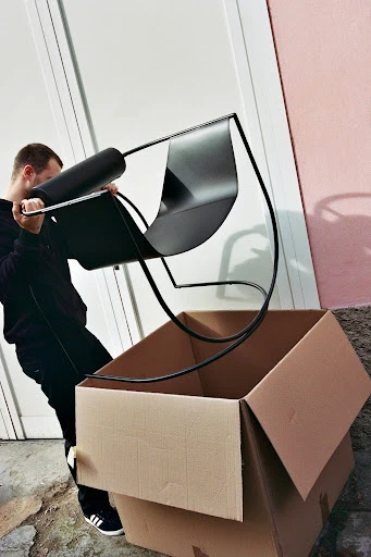 Person lifting a black modern chair to place it inside a large cardboard box next to a white door and pink wall outside.