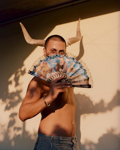 Man holding a decorative folding fan in front of his face with animal horns positioned behind his head by photographer Paolo Zerbini.