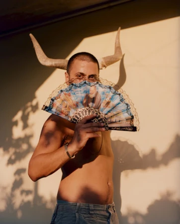 Man holding a decorative folding fan in front of his face with animal horns positioned behind his head by photographer Paolo Zerbini.