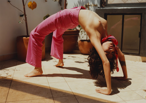 A woman in a backbend with pink pants and a red bandana poses on a tiled patio with sunlight and plants in the background.