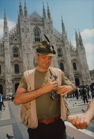 Man wearing a green feathered hat and utility vest holding pigeons in front of a grand cathedral exterior.