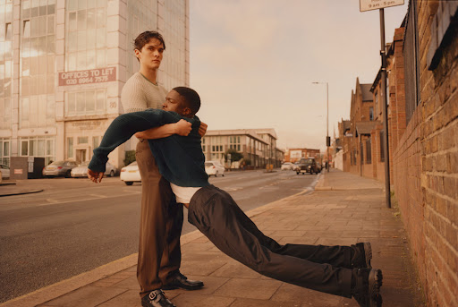 Two men on a city sidewalk: one African-American man is being held up by a Caucasian man wearing brown slacks and a cream sweater.