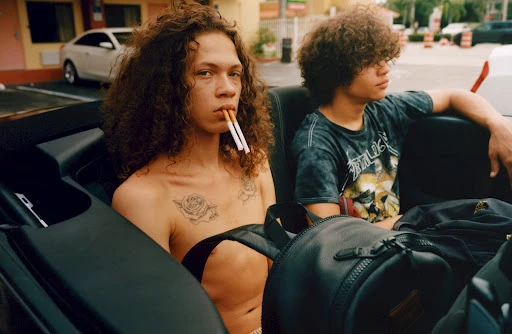 Two young men in a convertible car. One has cigarettes in his mouth. The other wears a band t-shirt and has curly hair.