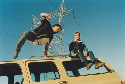 Two adult males sit on a tan SUV outdoors during the daytime; one sits while the other balances with a power line in the background.