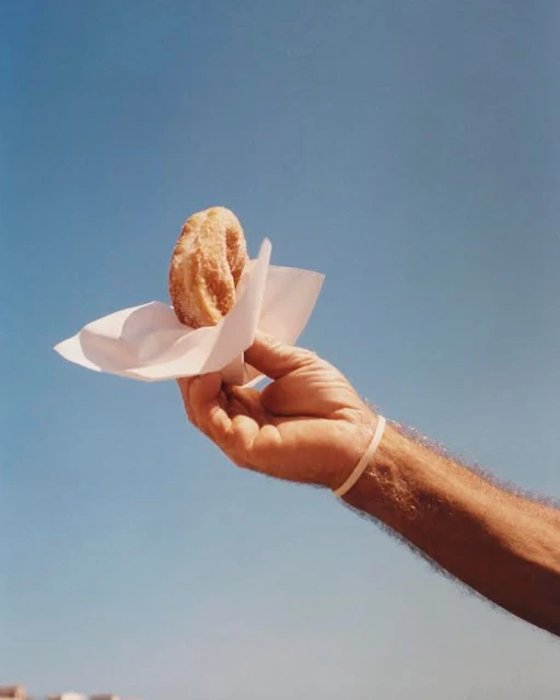 A hand holding a churro with a paper napkin against a clear blue sky, providing a simple, bright, and appetizing image.