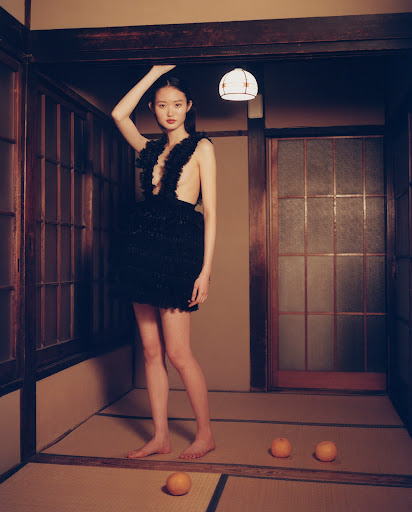 A young woman in a black dress poses indoors with fruit on the floor, showcasing fashion in an atmospheric, editorial style shot.