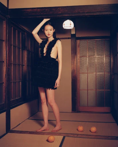 A young woman in a black dress poses indoors with fruit on the floor, showcasing fashion in an atmospheric, editorial style shot.