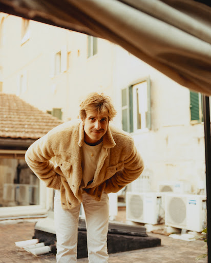 Full-length portrait of a smiling man standing on a rooftop wearing a beige jacket, white jeans, and a cream-colored sweater. 