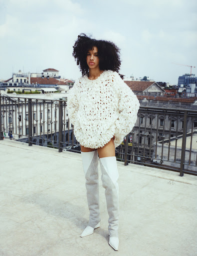 Fashionable woman posing in an oversized sweater and thigh-high boots on a rooftop overlooking a cityscape, showcasing style and urban sophistication.