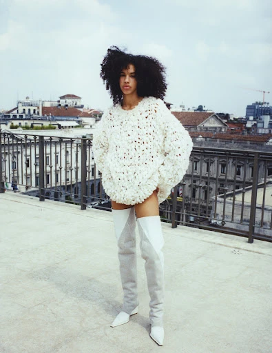 Fashionable woman posing in an oversized sweater and thigh-high boots on a rooftop overlooking a cityscape, showcasing style and urban sophistication.