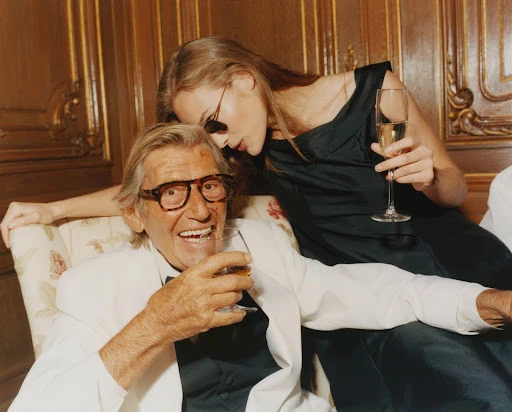 Elderly man in white suit smiles while holding glass, young woman in black dress stands behind him holding champagne flute.