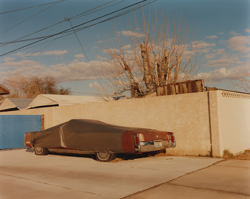 Classic maroon car parked on a concrete pad and covered with a dark tarp next to a tan stucco wall and fence on a sunny day.