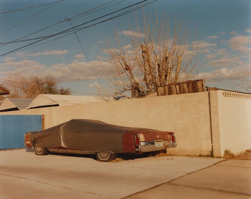 Classic maroon car parked on a concrete pad and covered with a dark tarp next to a tan stucco wall and fence on a sunny day.