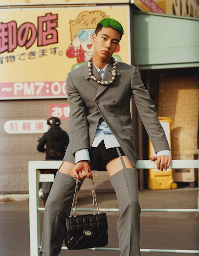 Young man in grey suit and green hair poses with handbag in urban street scene with Japanese signage and police officer in the background.